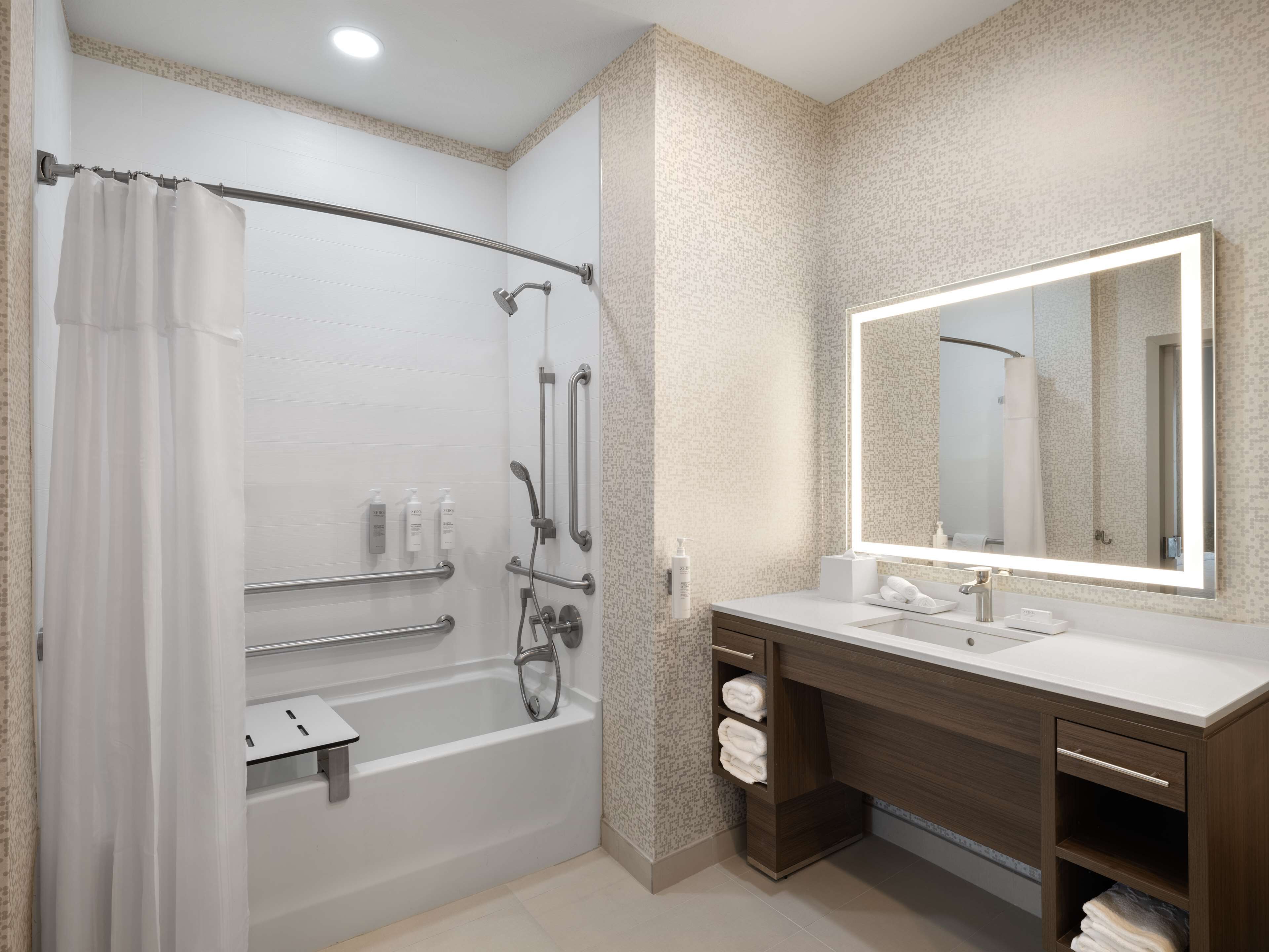 Un baño moderno con ducha a ras de suelo, barras de apoyo, un espejo grande y un lavabo con toallero.