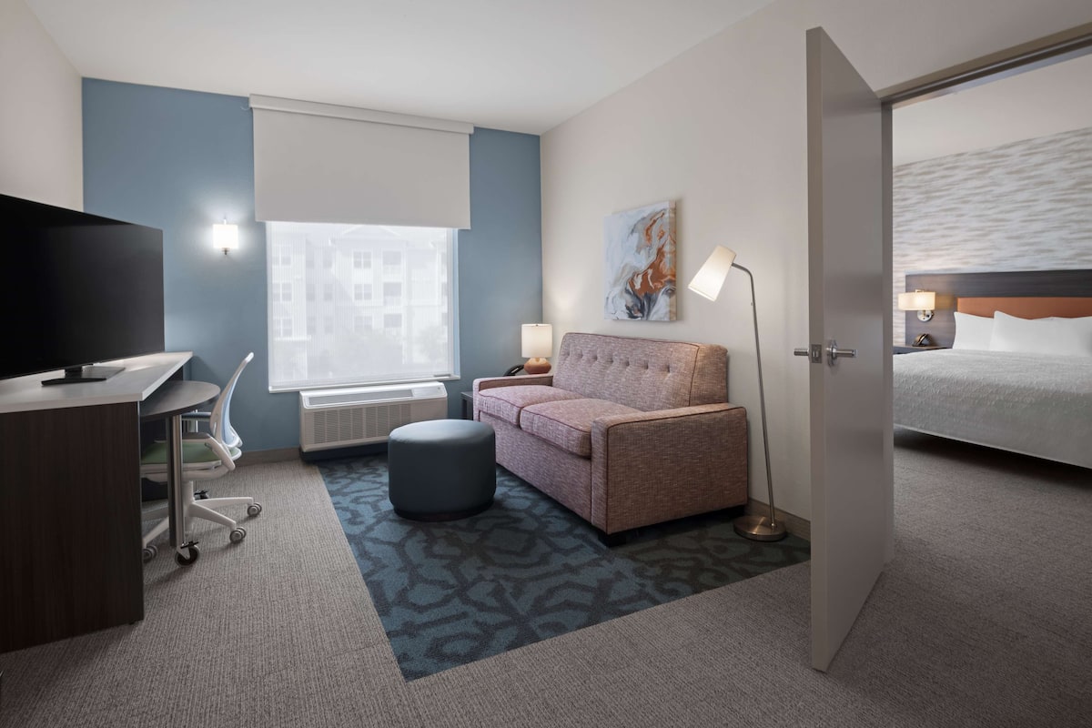 Ett hotellrum med en säng, en soffa, ett skrivbord med en tv, en lampa och ett fönster med persienner.