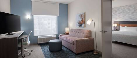 Habitación de hotel con cama, sofá, escritorio con televisor, lámpara y ventana con persianas.