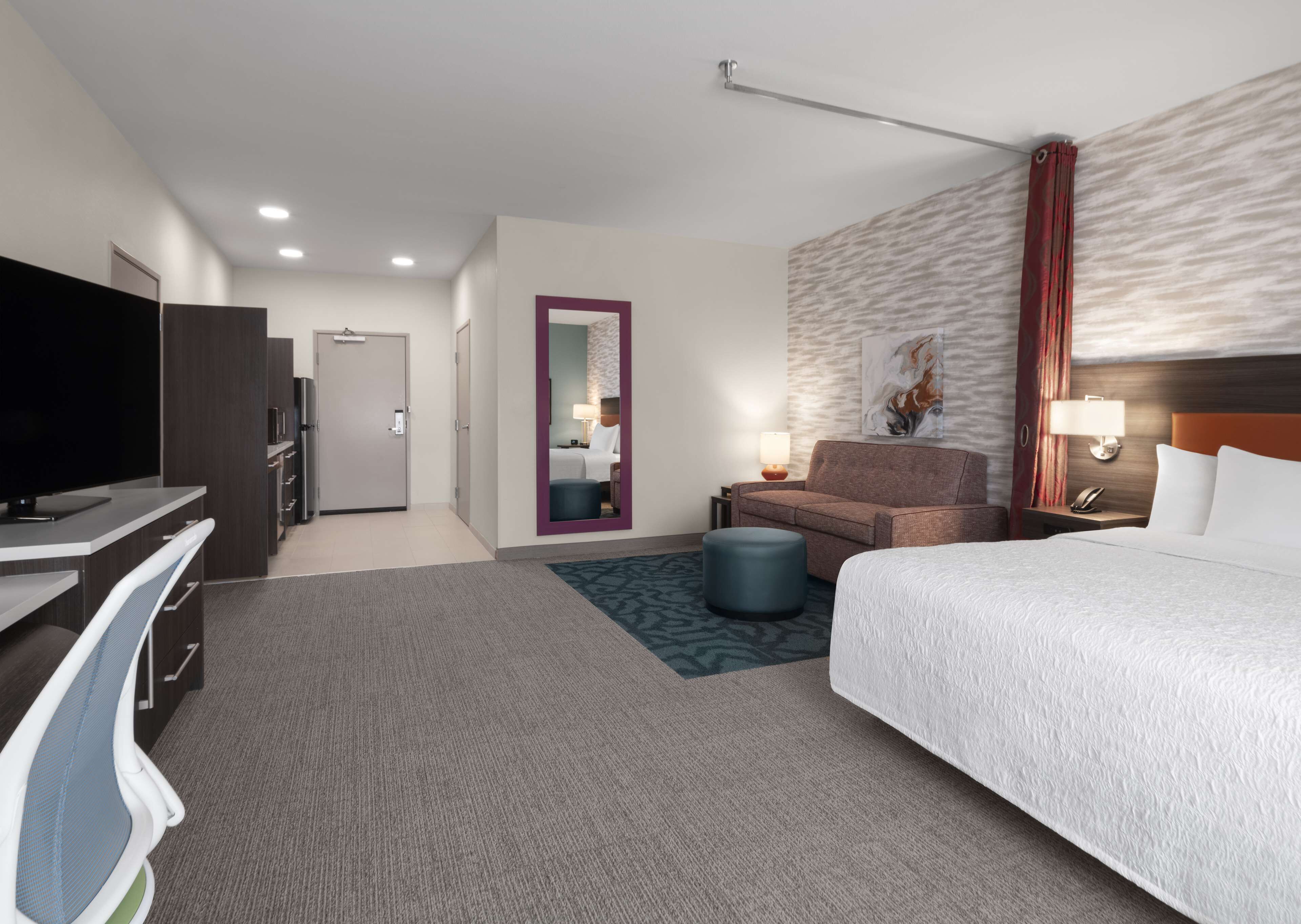 A hotel room with a bed, a desk with a TV, a sofa, a chair, a small table with a lamp, and a wall-mounted artwork.