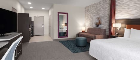 A hotel room with a bed, a desk with a TV, a sofa, a chair, a small table with a lamp, and a wall-mounted artwork.