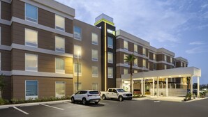 Un hotel de varios pisos con un diseño moderno, que destaca un letrero grande de 'HOME2', un acceso cubierto y un área de estacionamiento con varios vehículos.
