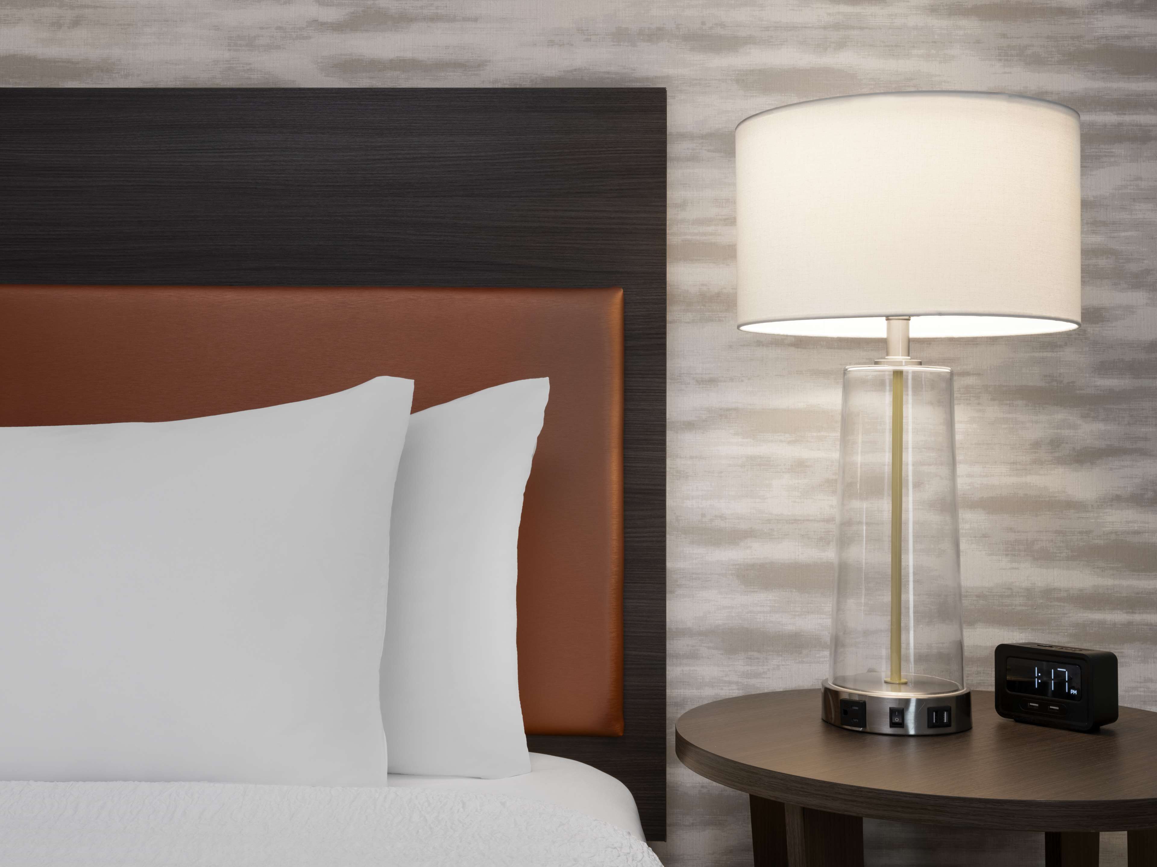 Et hotelværelse med en seng, et natbord, en lampe og et digitalt ur.