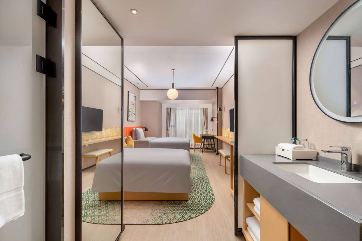 A modern hotel room with a large bed, a bedside table, a round mirror, and a bathroom with a sink and mirror.