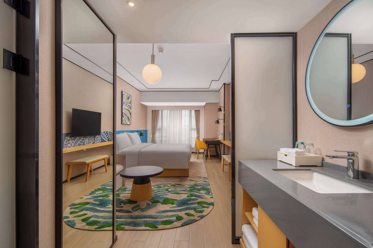 A modern hotel room with a bed, a desk, a round table, a large mirror, a TV, and a bathroom with a sink and mirror.