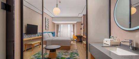 A modern hotel room with a bed, a desk, a round table, a large mirror, a TV, and a bathroom with a sink and mirror.