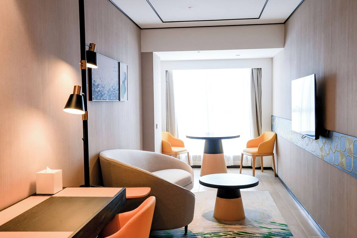 A modern hotel room with a curved sofa, a round table, and two chairs. There is a flat-screen TV mounted on the wall and a large window with curtains.