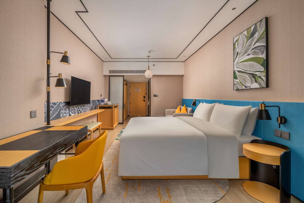 A hotel room with a bed, desk, chairs, a TV, and a painting on the wall.