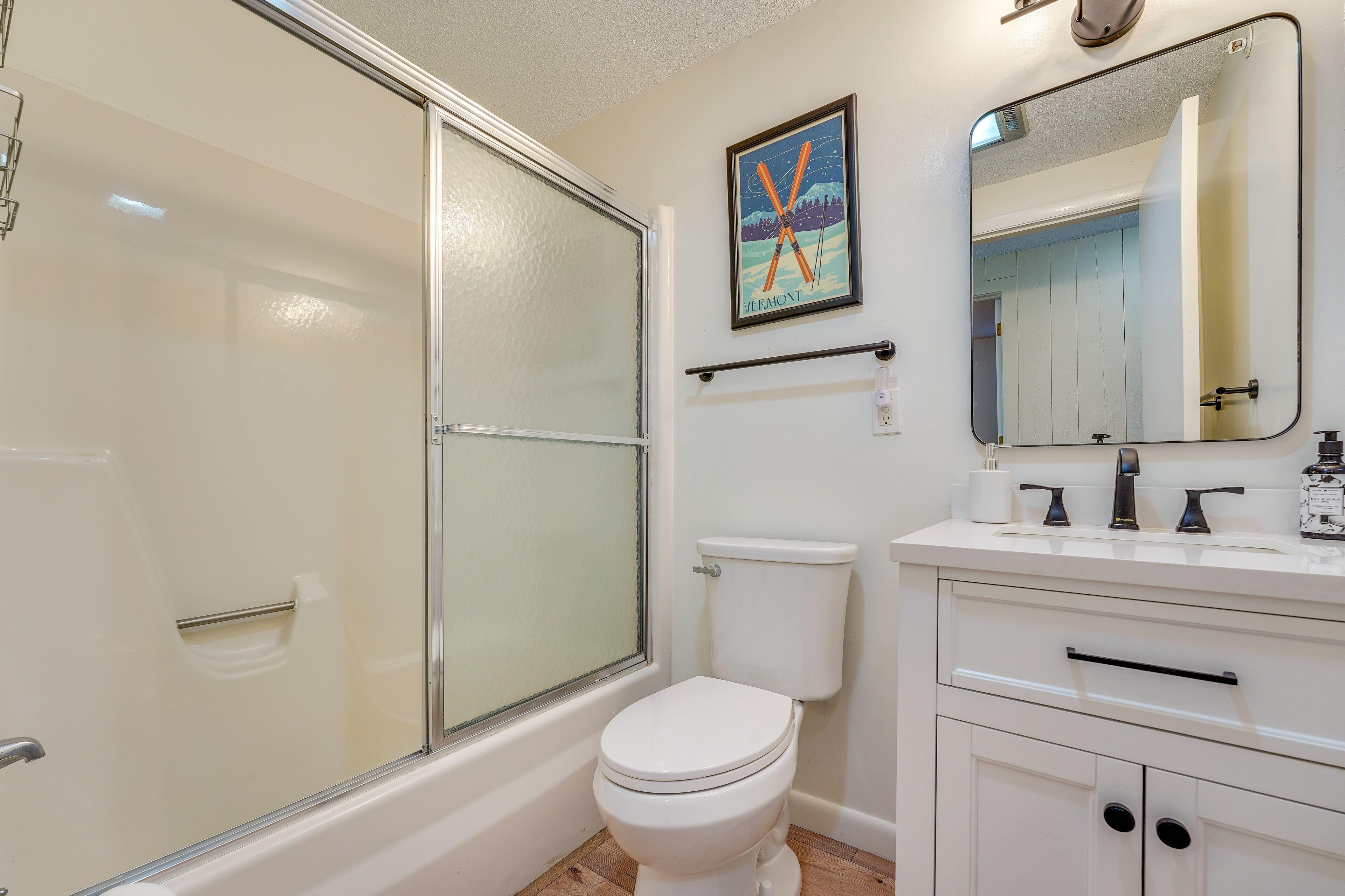 A bathroom with a glass shower enclosure, a white toilet, a sink with a mirror, and a framed picture on the wall.