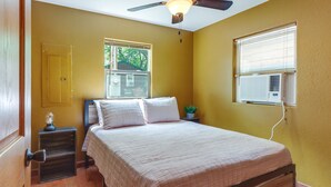 Un dormitorio con cama, mesita de noche, ventilador de techo, ventana con persianas y aire acondicionado.