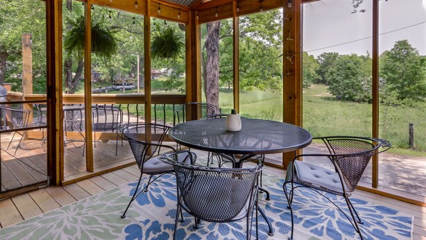Un patio techado al aire libre con una mesa redonda y sillas, rodeado de amplios ventanales con vistas a un paisaje verde.
