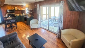 Una sala de estar acogedora con un sofá, una mesa de centro y un televisor. Hay una zona de cocina con microondas y una mesa de comedor. La habitación tiene pisos de madera y un ventilador de techo.