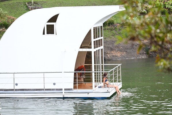 Una casa flotante con una cubierta amplia y abierta y un ventanal pequeño, ubicada sobre aguas tranquilas.