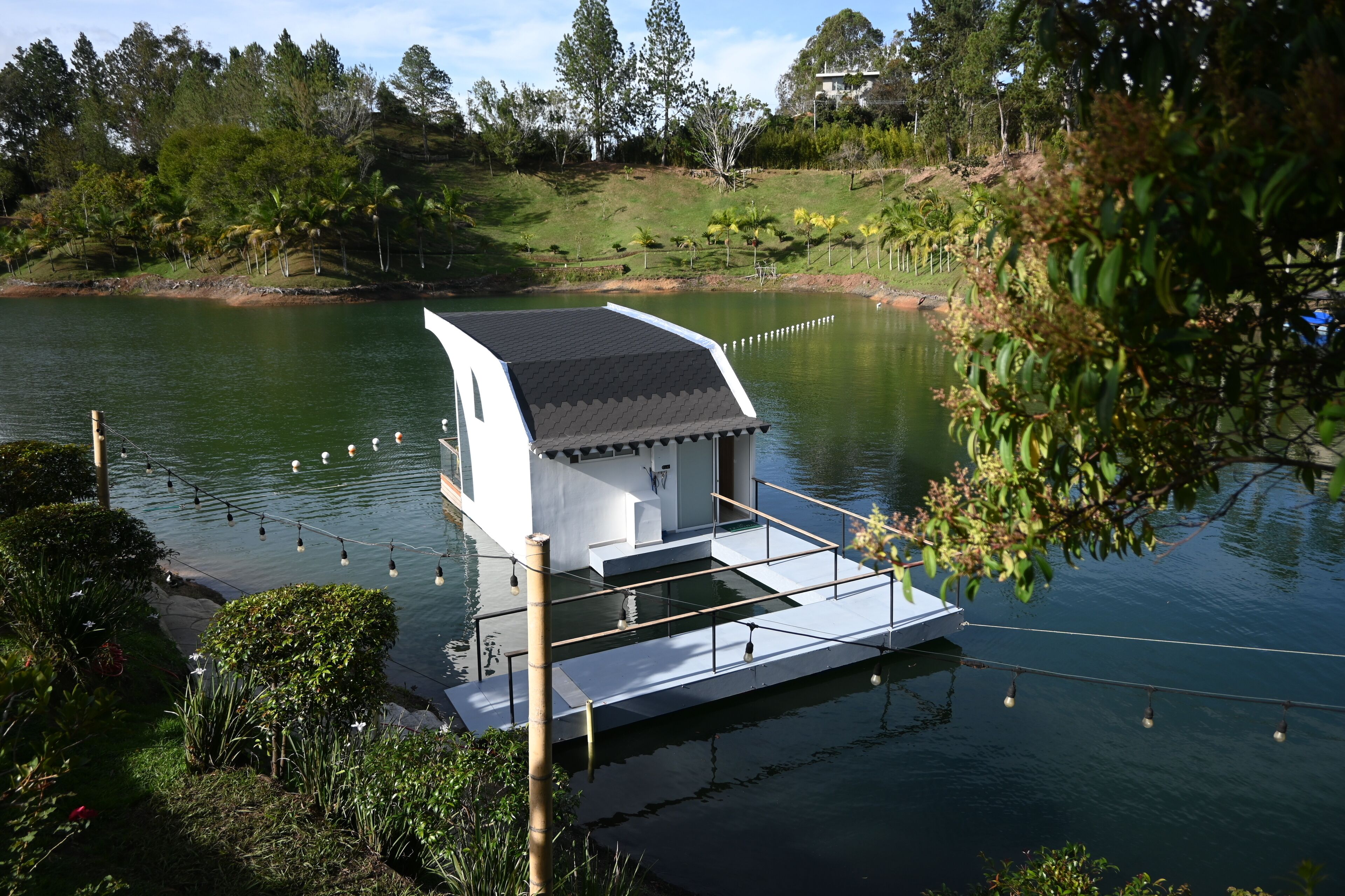 Una casa flotante con techo plano y balcón, amarrada en un lago tranquilo rodeado de vegetación.