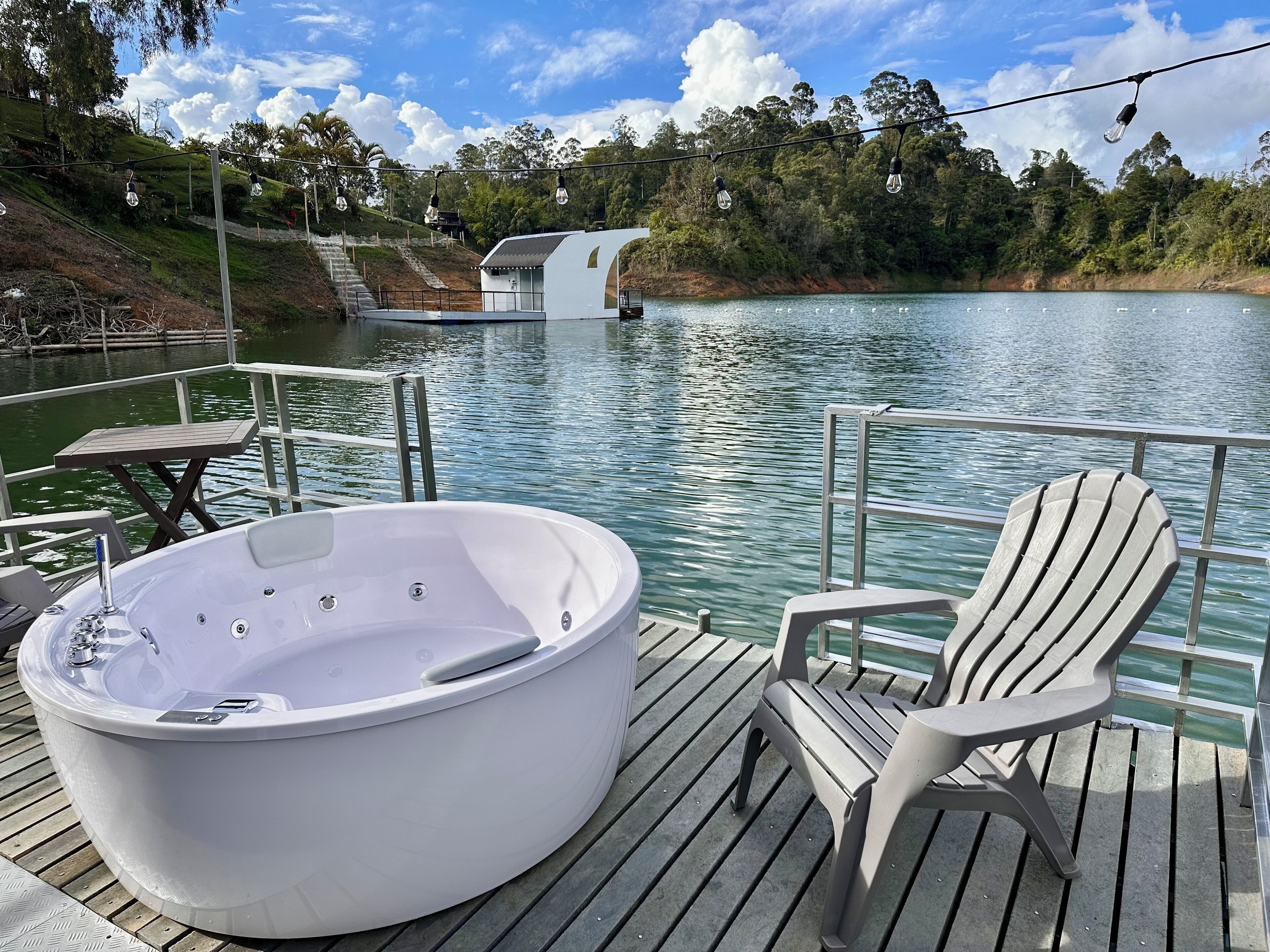 A hot tub on a wooden deck with a chair, overlooking a lake with a house in the background.