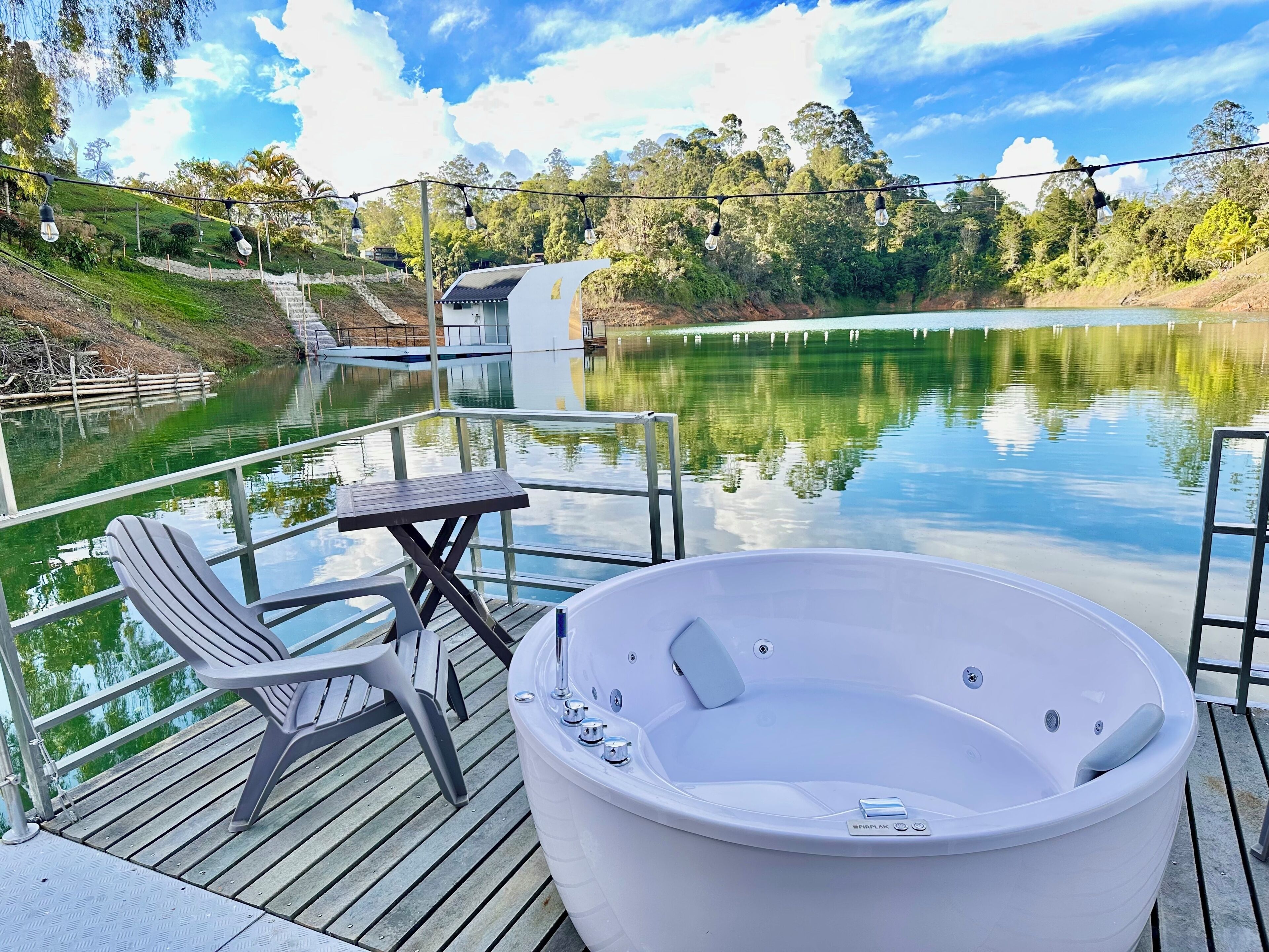 Une terrasse dotée d’un jacuzzi, d’une table et de chaises, offrant une vue sur un lac, avec un quai et une végétation luxuriante en arrière-plan.