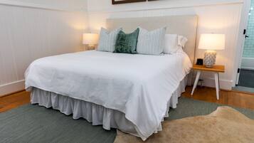 A neatly made bed with a white bedspread and multiple pillows, a wooden nightstand with a lamp, and a green and beige rug on a wooden floor.