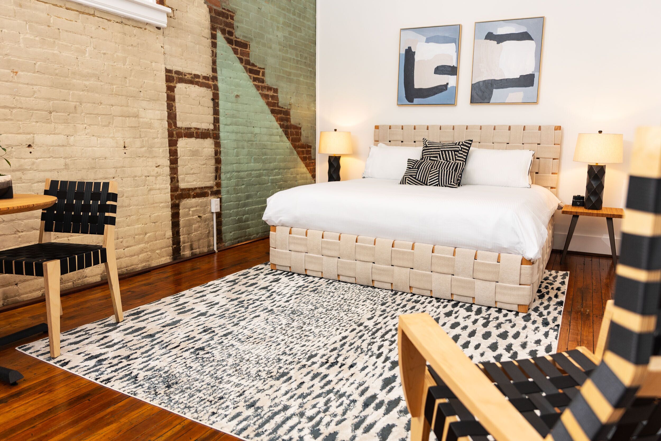 A hotel room with a brick wall, a bed with a white headboard, two bedside tables with lamps, a patterned rug, and a wooden chair.