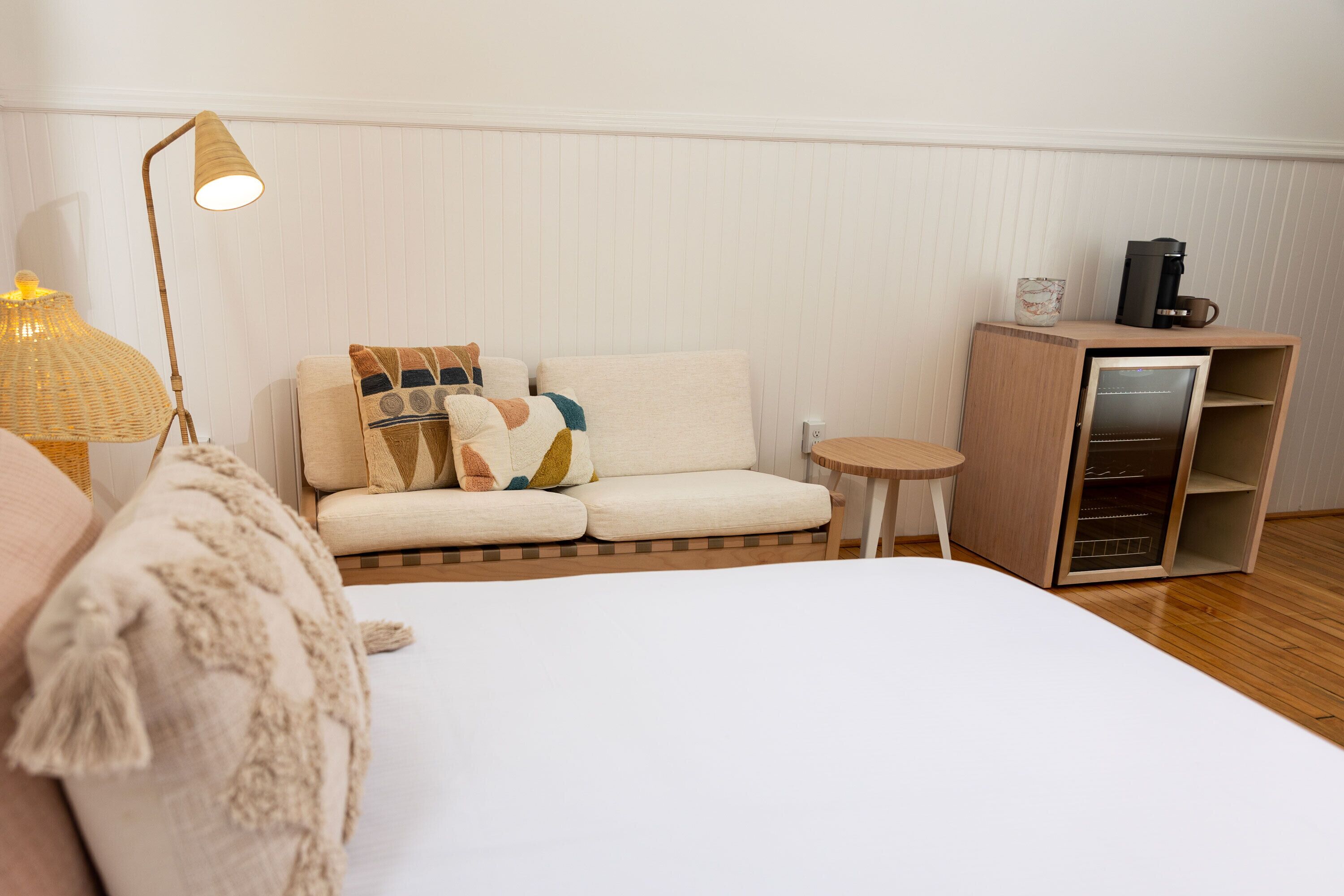 A neatly arranged hotel room with a bed, a sofa, a small table, a lamp, and a coffee machine.