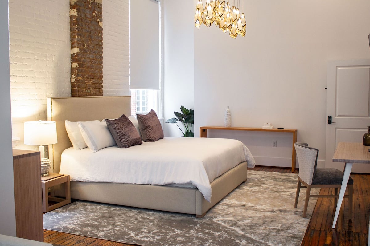 Ein Schlafzimmer mit einem großen Bett, Nachttischen, einem kleinen Tischchen, einer hölzernen Bank, einem Sessel, einer Zimmerpflanze und einer Pendelleuchte.