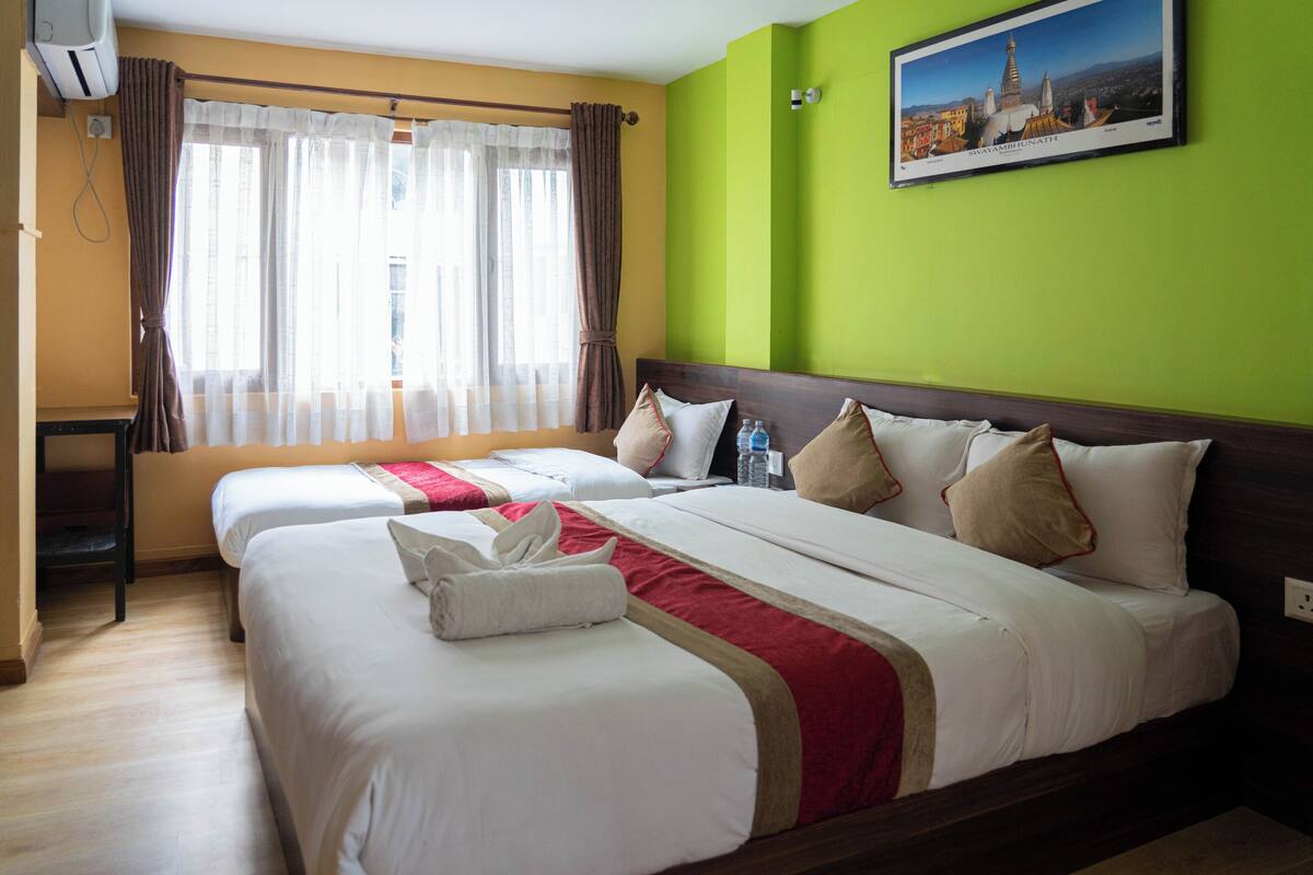 A hotel room with two beds, a large window with curtains, a green wall, and a framed picture.