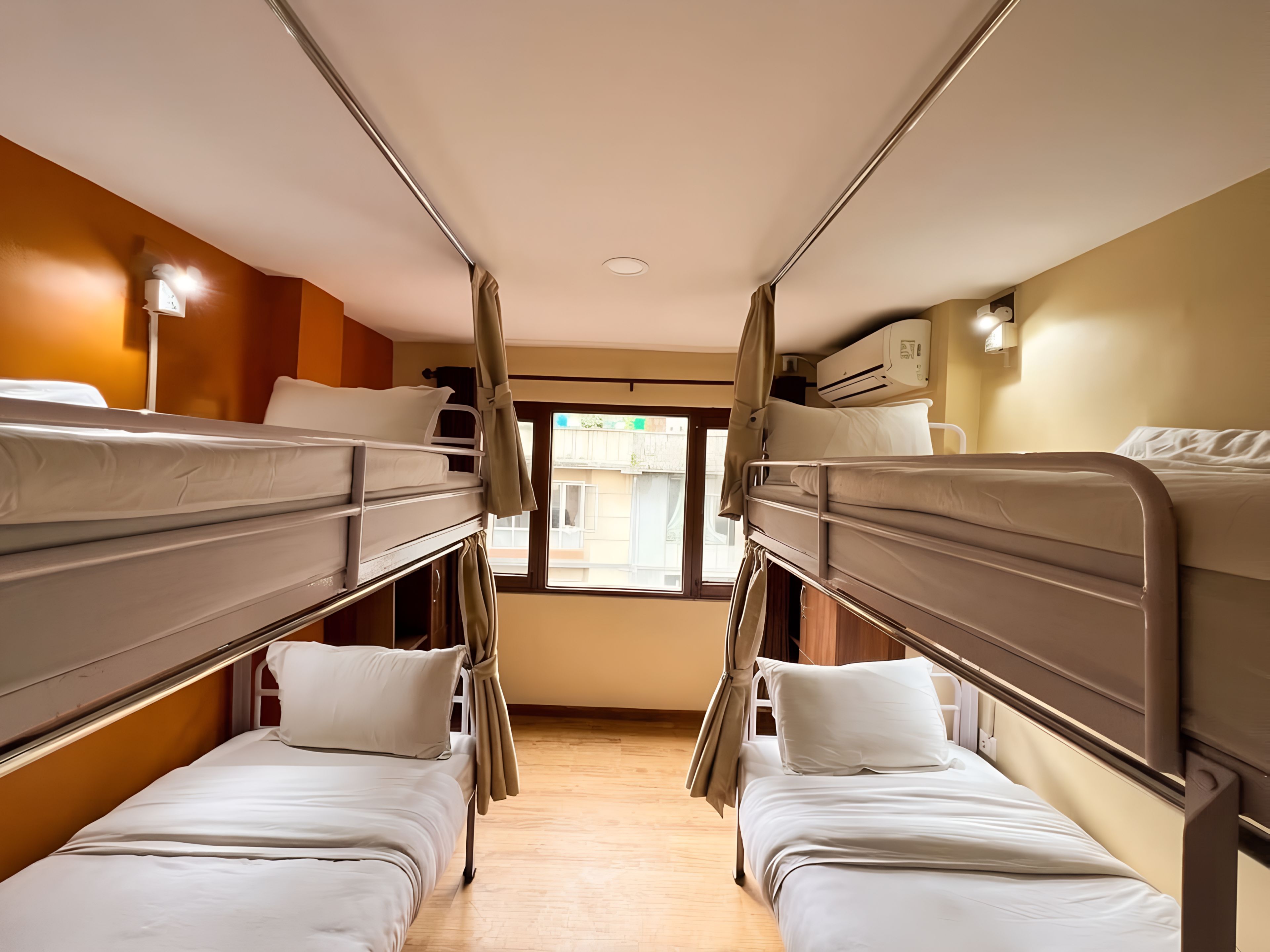 A train compartment with two bunks, each with a mattress and pillows, a window with a view of the outside, and an air conditioning unit on the wall.