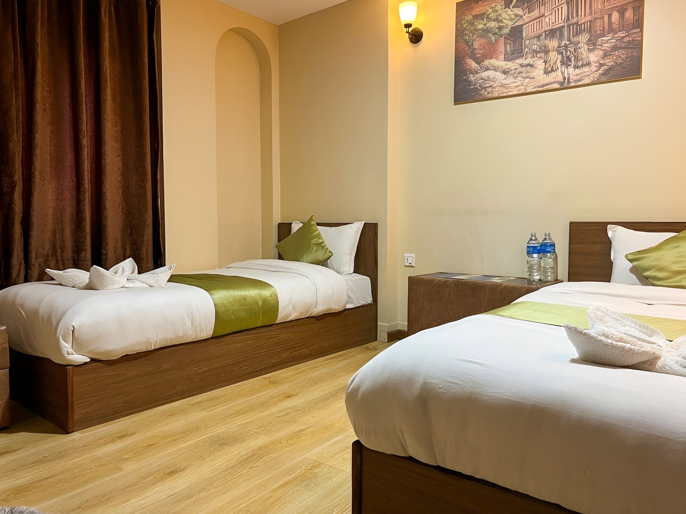 A hotel room with two beds, wooden headboards, a painting on the wall, and a nightstand with water bottles.