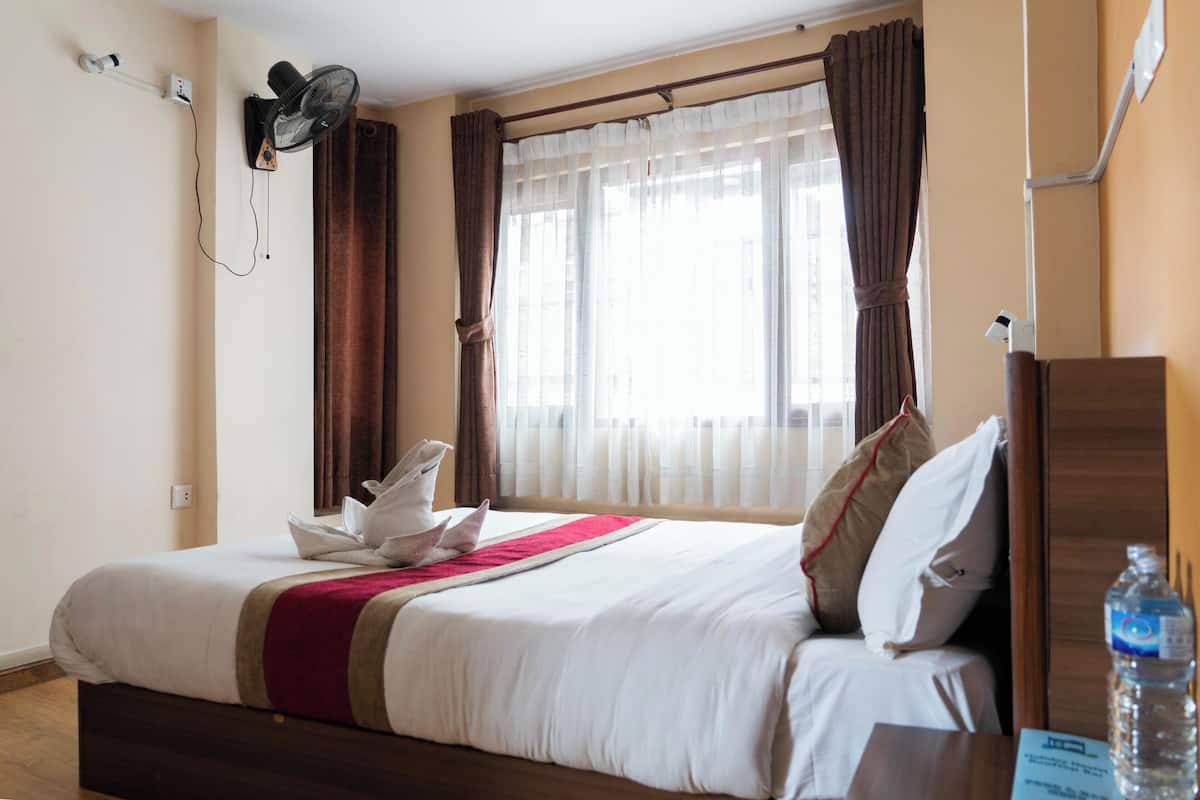 A hotel room with a bed, a wall-mounted fan, a window with curtains, and a water bottle on the nightstand.