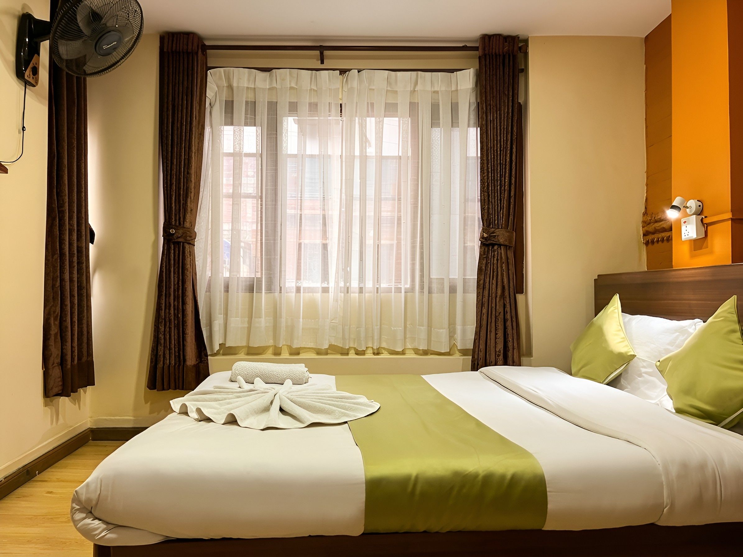 A hotel room with a bed, a window with curtains, a wall-mounted fan, and a nightstand with a lamp.