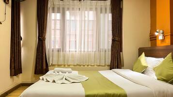 A hotel room with a bed, a window with curtains, a wall-mounted fan, and a nightstand with a lamp.