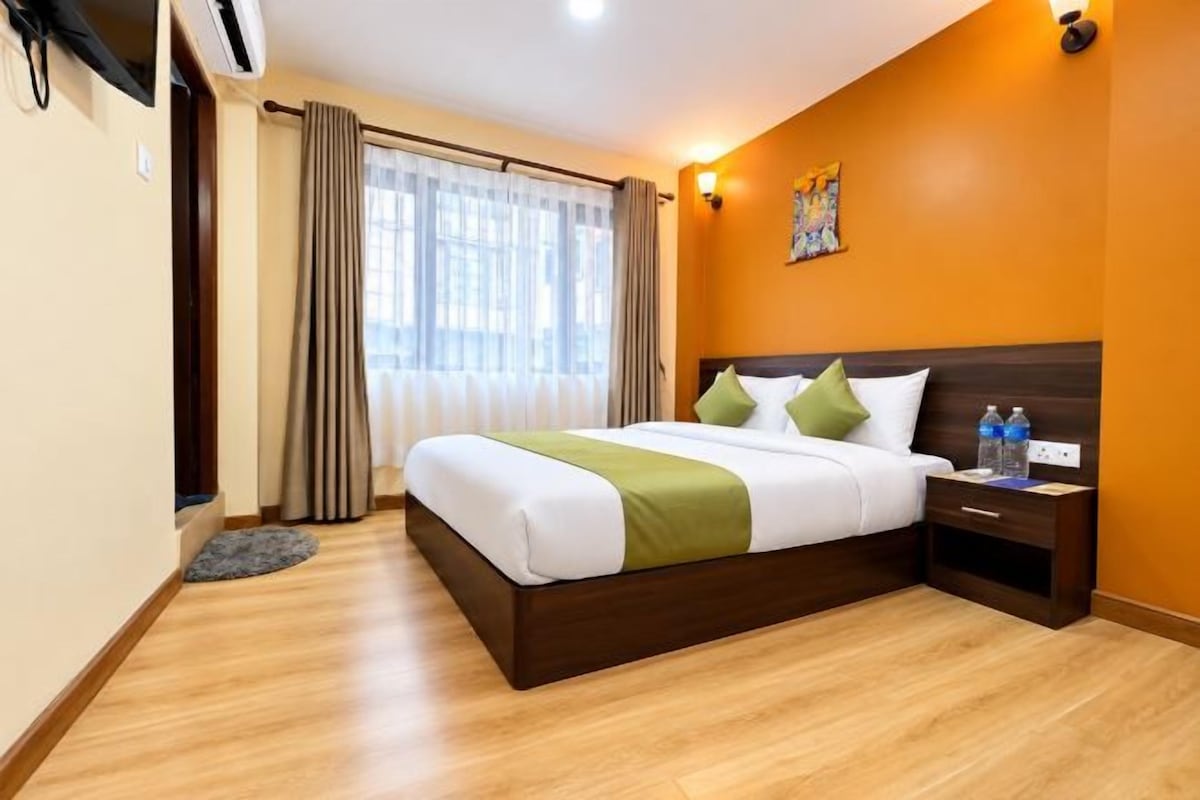 A hotel room with a bed, a nightstand, a window with curtains, and a wall-mounted light fixture.
