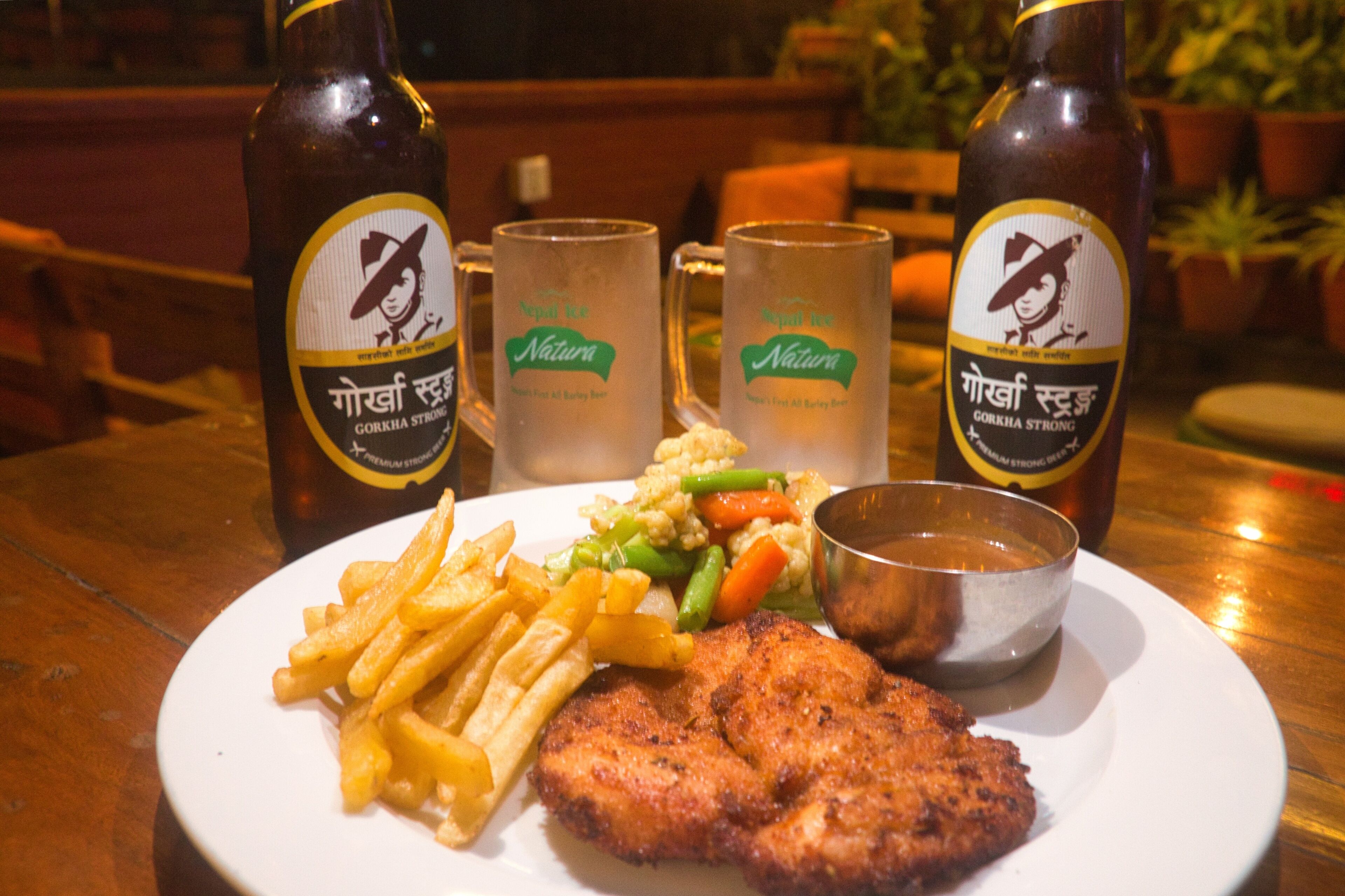 A plate with a meat dish, fries, and vegetables, accompanied by two beer bottles and two mugs on a wooden table.