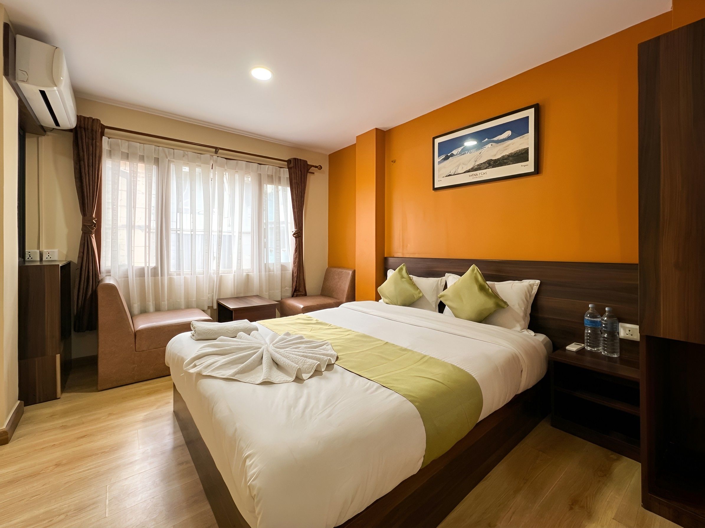 A hotel room with a bed, a chair, a desk, an air conditioning unit, and a painting on the wall.