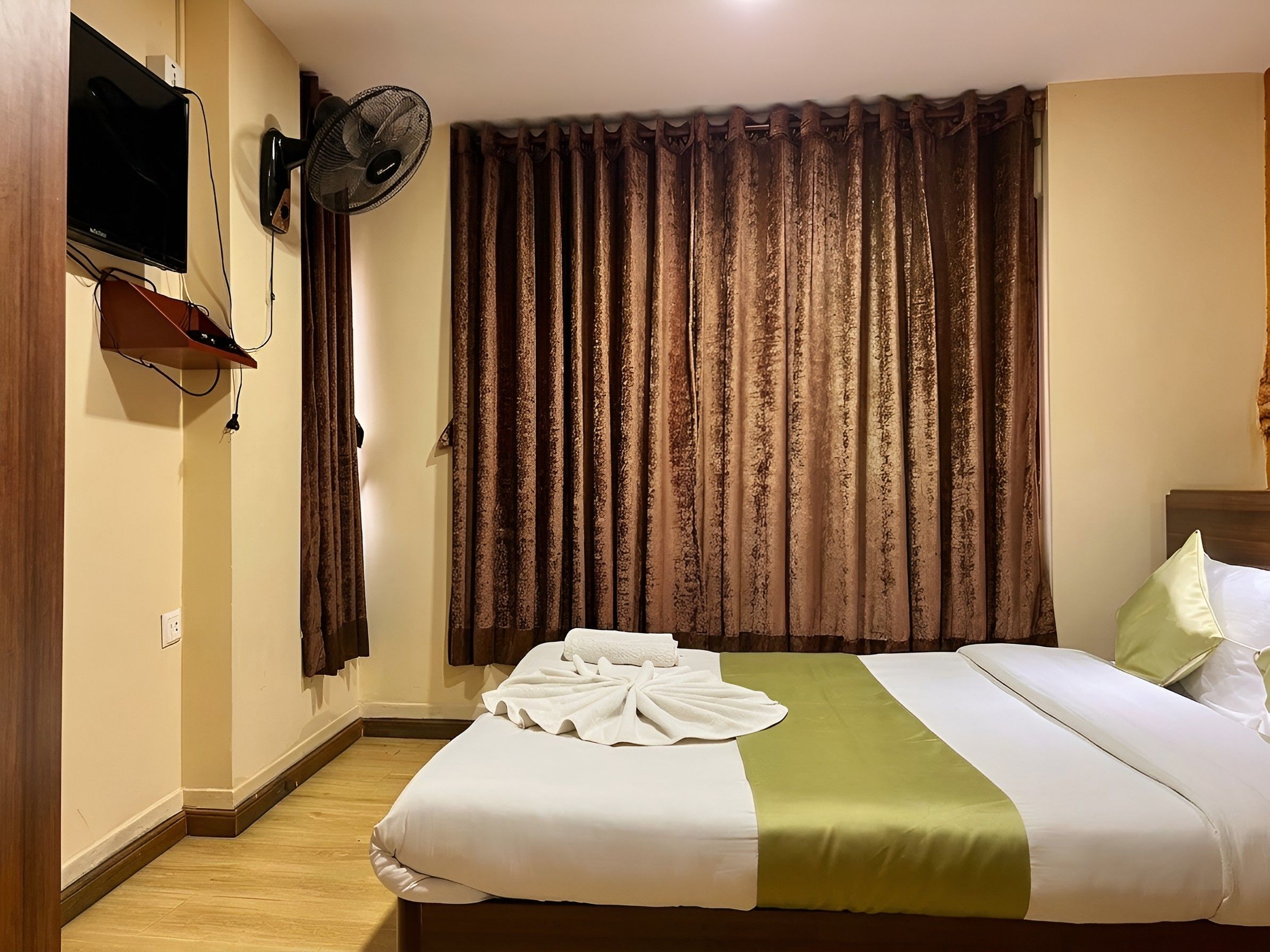 A hotel room with a bed, a flat-screen TV mounted on the wall, a standing fan, and a window with curtains.