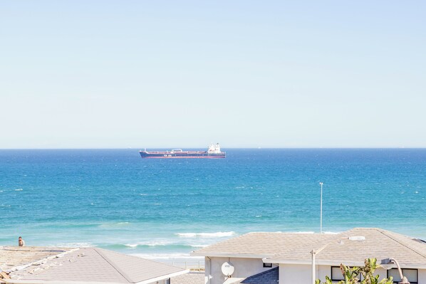 A coastal view with a large cargo ship in the distance, a beach, and a building with a satellite dish. - Beachfront couples paradise overlooking Robben Island and Atlantic ocean. (Cape Town)