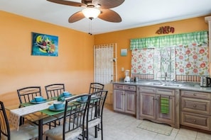 Une cuisine avec une table à manger, des chaises, un ventilateur de plafond et une fenêtre agrémentée de rideaux à motifs floraux.