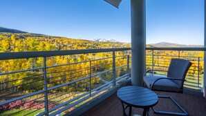 Ein Balkon mit Metallgeländer, einem Tisch und einem Stuhl mit Blick auf eine bewaldete Landschaft und Berge in der Ferne.