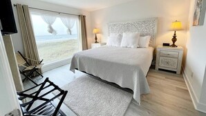 Uma cama de casal com cabeceira estampada, duas luminárias de cabeceira, uma cadeira e vista para a praia através de uma janela ampla.