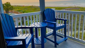 Uma varanda com cadeiras Adirondack azuis e uma pequena mesa, com vista para a praia e o oceano.