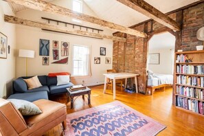 A cozy living room with a sofa, a coffee table, a rug, a bookshelf, and a dining area with a table and chairs. - The Railway Bridge Cottage (Catskill)