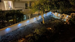 Vue nocturne d’un jardin aménagé, avec un mur blanc, des arbres et des lumières illuminées.