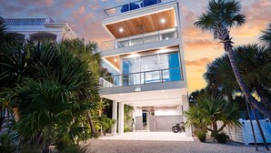 A modern multi-story house with a garage, surrounded by tropical vegetation. - #1 Rooftop Deck w/ Gulf Views - Steps to Beach (Siesta Key)