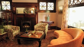 A cozy living room with a fireplace, floral-patterned armchairs, a coffee table with books and a chess set, and a view through a window with patterned curtains. - Marianna Stoltz House (Spokane)