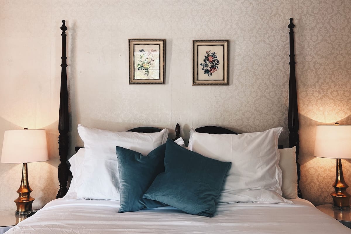 A neatly made bed with white and teal pillows, flanked by two wall-mounted picture frames, and bedside lamps on either side.