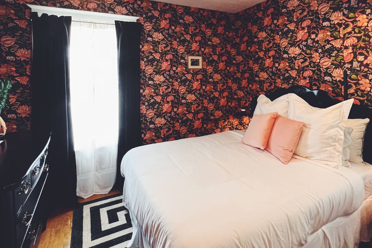 A bedroom with a bed, a desk, and a chair. There is a window with curtains and a lamp hanging from the ceiling.