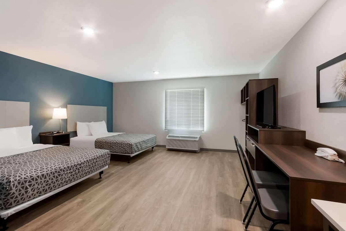 A hotel room with two beds, a desk, a chair, a TV, a lamp, and a window with blinds.