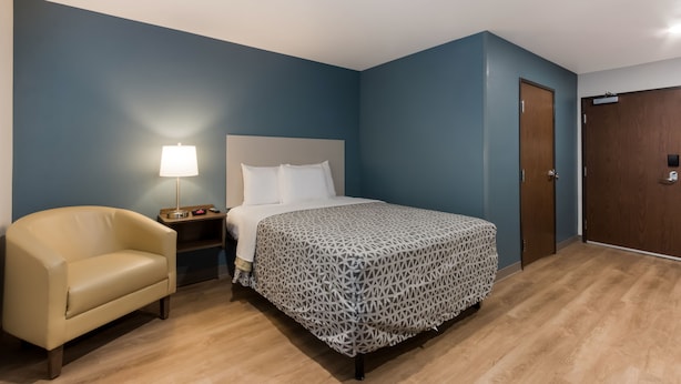 A bedroom with a bed, a nightstand, a lamp, a chair, and a wardrobe. - Woodspring Suites Chandler Airport (Chandler)