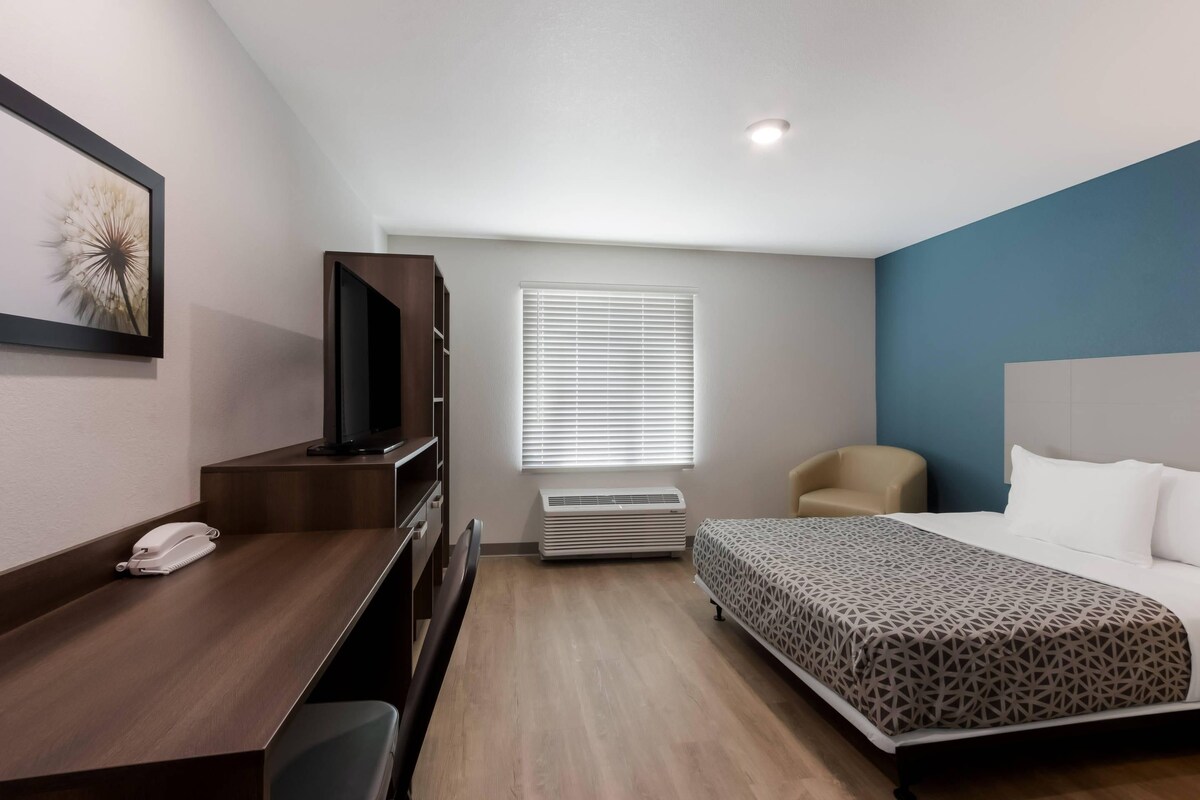 A hotel room with a bed, a desk with a telephone, a chair, a television, a window with blinds, and an air conditioning unit.