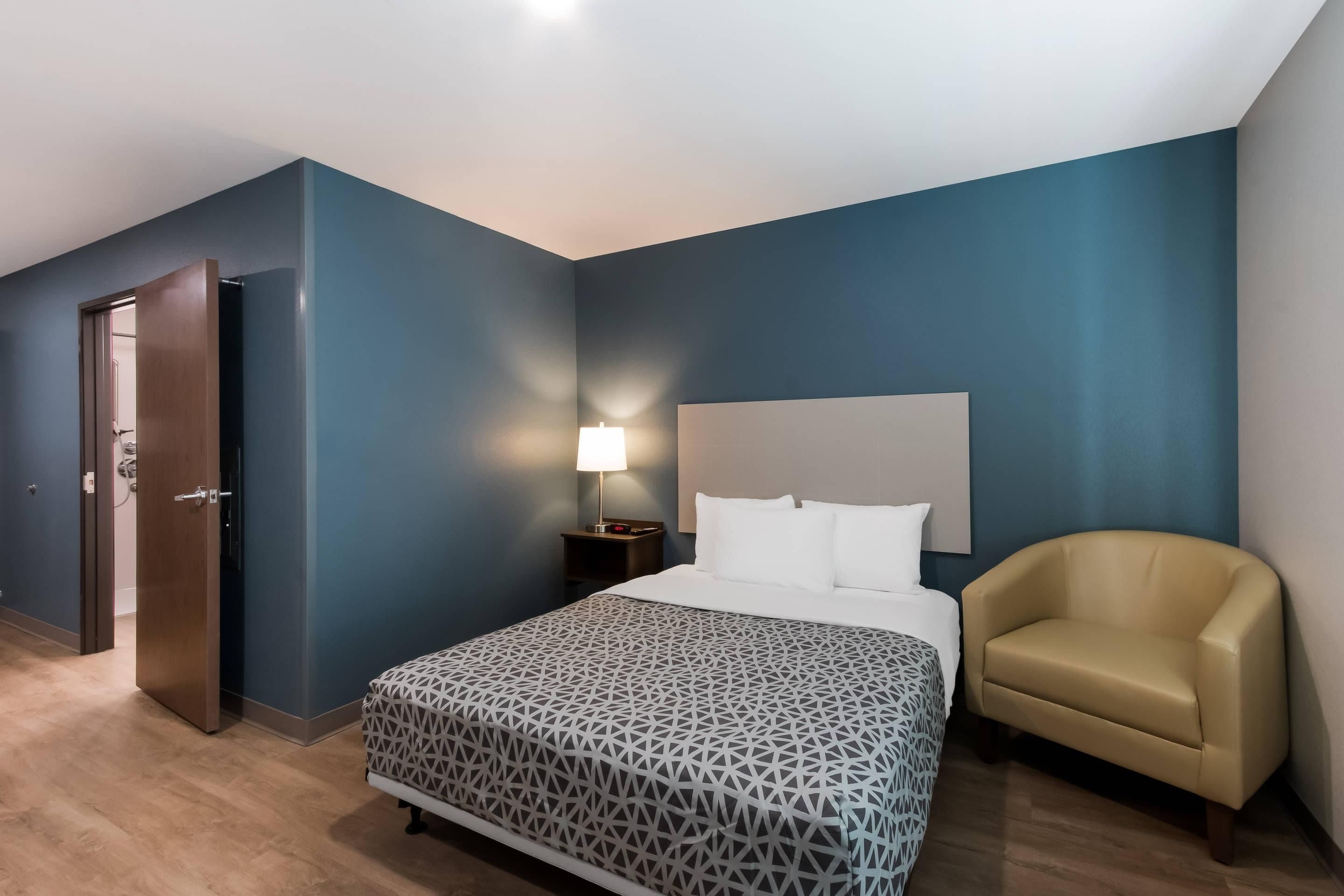 A hotel room with a bed, a chair, a nightstand with a lamp, and a door.