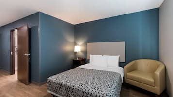 A hotel room with a bed, a chair, a nightstand with a lamp, and a door.